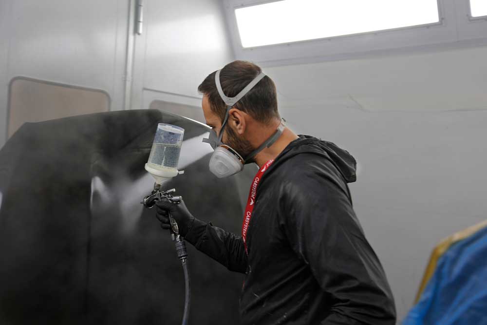 Fahrzeug- und Kleinserienlackierung-Mitarbeiter lackiert im Stehen mit dem Behältnis, das an einer Pistole angebracht ist, eine schwarze Motorhaube