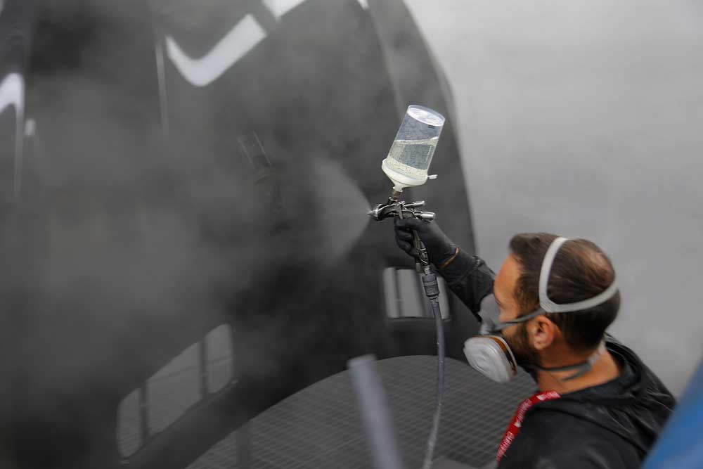 Fahrzeug- und Kleinserienlackierung-Mitarbeiter lackiert im Knien mit dem Behältnis, das an einer Pistole angebracht ist, eine schwarze Motorhaube