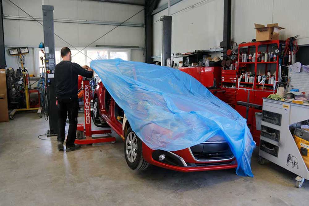 Aufbereitung von Leasingrückläufern-Mitarbeiter fängt an, ein rotes Auto mit einer blauen Folie abzudecken, im Hintergrund ist das Lackierwerk Jakobson PKW zu sehen