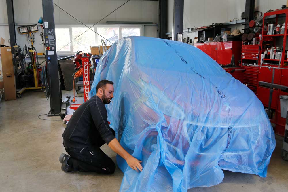 Aufbereitung von Leasingrückläufern-Mitarbeiter deckt ein rotes Auto mit einer blauen Folie ab, im Hintergrund ist das Lackierwerk Jakobson PKW zu sehen