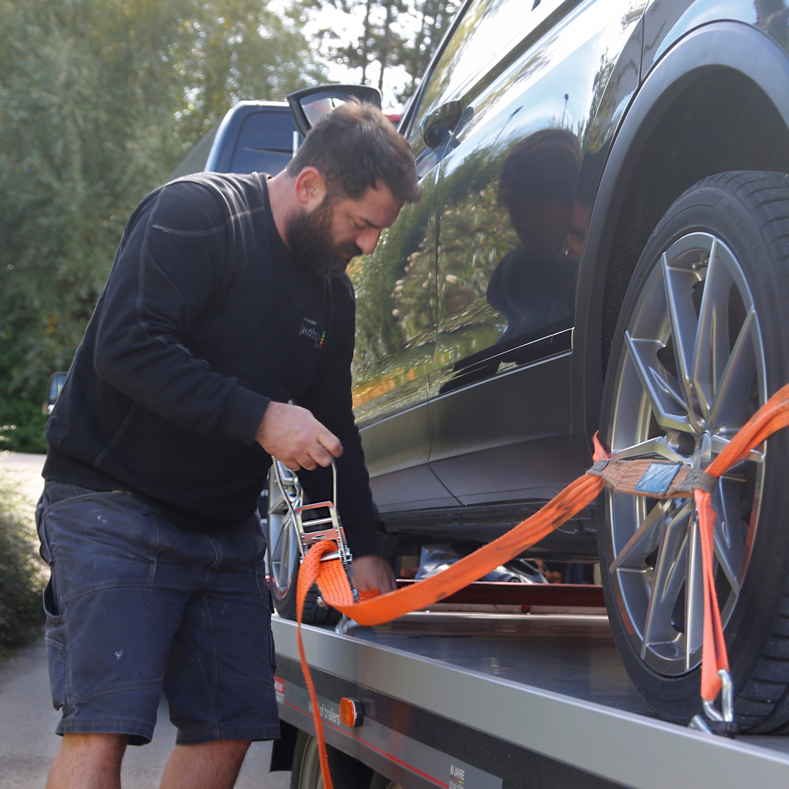 jakobson-services-abhol-bring-service Services-Kostenloser Abhol- und Bringservice-Der Abteilungsleiter befestigt mit einem orangenen Spanngurt die Reifen eines grauen Autos auf einem Anhänger
